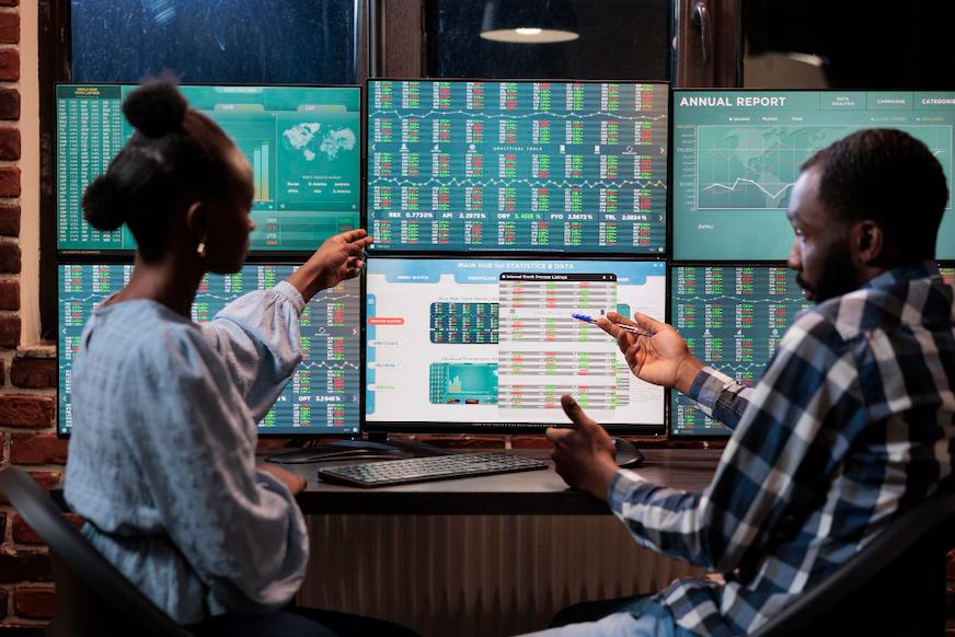 Two engineers pointing at a wall of monitors showing live dashboards.
